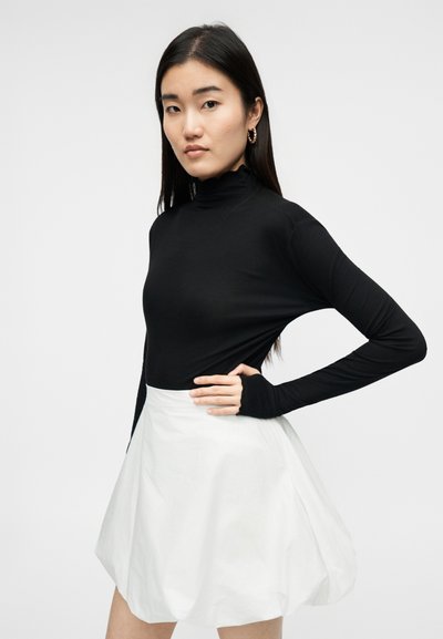 Νεαρή γυναίκα με μακριά σκούρα μαλλιά, φοράει χρυσούς κρίκους στα αυτιά, μαύρο ζιβάγκο με μακριά μανίκια και λευκή φούστα με άνοιγμα, ποζάροντας με το ένα χέρι στη μέση.