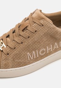 Beige Wildleder-Sneaker mit perforiertem Material, tonal passenden Schnürsenkeln und geprägtem Logo-Detail. Die Sohle ist weiß und hat eine strukturierte Oberfläche.