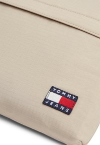 Μπεζ θήκη λάπτοπ από υφασμάτινη υφή. Διαθέτει ορθογώνιο σχήμα και κεντημένο λογότυπο Tommy Jeans σε μπλε, κόκκινο και άσπρο.