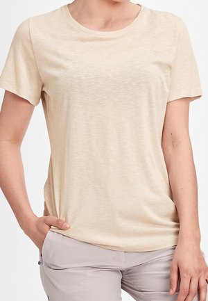 Detail osoby, která má na sobě béžové tričko s krátkým rukávem a světle šedé kalhoty, s jednou rukou v kapse, na jednobarevném pozadí.