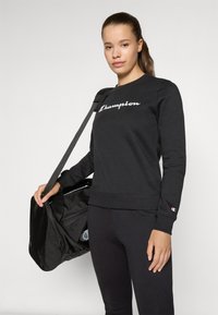 Zwarte sweatshirt met wit "Champion" logo, ronde halslijn, geribbelde mouwen, gecombineerd met zwarte broek en een zwarte tas.