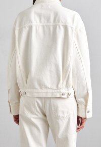 Bílá džínová bunda s volným střihem, knoflíkovými detaily a bočními kapsami. Má límec a viditelné švy na strukturované látce.