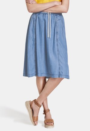 Vrouw draagt een lichtblauwe denimrok tot op de knie met een wit trekkoord, gekruiste armen en bruine gevlochten sandalen met enkelbandje.
