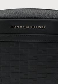 Musta teksturoitu nahkareppu, jossa on kohokuvioitu neliökuvio ja metallinen "Tommy Hilfiger" -logo ylemmässä keskiosassa.