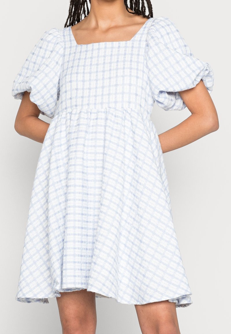 Femme portant une robe à carreaux blanche et bleu clair avec des manches courtes bouffantes et un col carré, debout les mains sur les hanches.