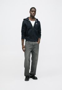 Tengerészkék cipzáras kapucnis pulóver kenguruzsebbel, világosszürke, egyenes szárú farmerrel és fekete cipővel. A modell fehér háttér előtt áll.