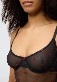 Μαύρο διαφανές εσώρουχο με κέντημα από κόκκινο κεράσι, με λεπτές τιράντες, κυματιστές άκρες και μια μικρή κόκκινη λεπτομέρεια κουμπιού στο κέντρο.