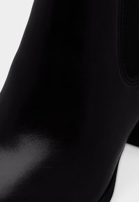 Gros plan sur une bottine en cuir noir brillant avec un panneau latéral élastique et un talon bloc sur fond blanc.