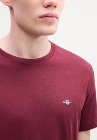 Close-up van een persoon die een maroon T-shirt met ronde hals draagt, met een klein geborduurd "GANT" logo op de linkerborst.