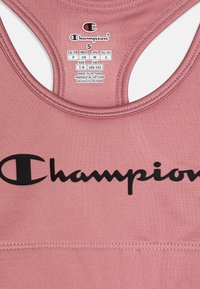 Rosa idrottstopp med ärmlös design, med en svart "Champion"-logga på framsidan, tillverkad av stretchigt material. Tvättråd finns inkluderat.