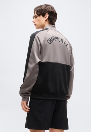 Νεαρός άνδρας φορώντας γκρι και μαύρο μπουφάν Champion F.C. και μαύρο σορτς, σε προφίλ, με απλό φόντο.