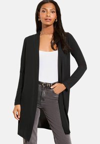 Fekete, hosszú kardigán puha anyagból, nyitott elülső résszel és hosszú ujjal. Fehér felsőre és szürke farmerre viselve.