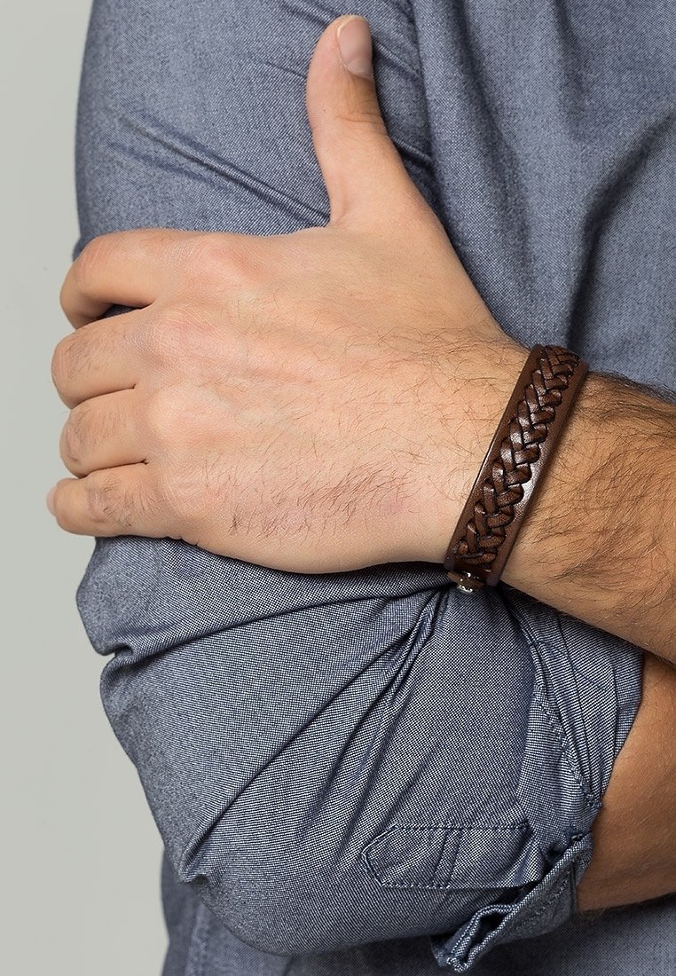 Bras adulte croisé sur la poitrine, portant une chemise bleue à manches longues et un bracelet en cuir tressé marron au poignet.