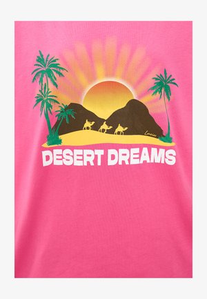 T-shirt en coton rose avec un design graphique représentant un soleil, des montagnes, des chameaux et des palmiers. Texte "DESERT DREAMS" en police blanche en gras.