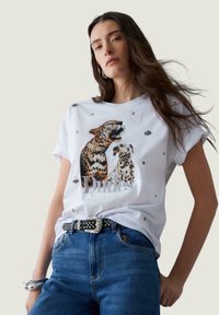 Bijela pamučna majica s tiskanim ilustracijom leoparda ukrašenom perlicama i natpisom "Dudes"; kombinirana s plavim trapericama i crnim remenom sa zakovicama.