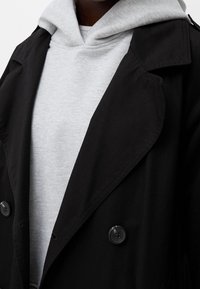 Czarny podwójnie zapięty płaszcz z teksturowanej tkaniny, z wysokim kołnierzem i dużymi czarnymi guzikami, noszony na jasnoszarym hoodie.