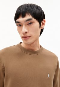 Jonge man met kort zwart haar, gekleed in een bruine ronde hals sweatshirt met een klein wit sleuteltje logo op de borst.