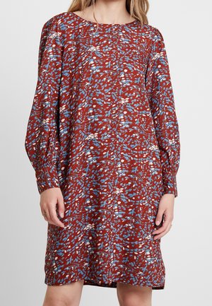 Mujer que lleva un vestido de manga larga hasta la rodilla con un fondo color óxido y un estampado floral disperso en azul y blanco.