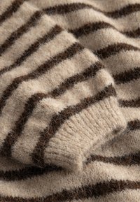 Material tricotat moale, bej, cu dungi orizontale maro închis și detaliu de manșetă cu nervuri, prezentând un prim-plan al unui pulover texturat și confortabil.