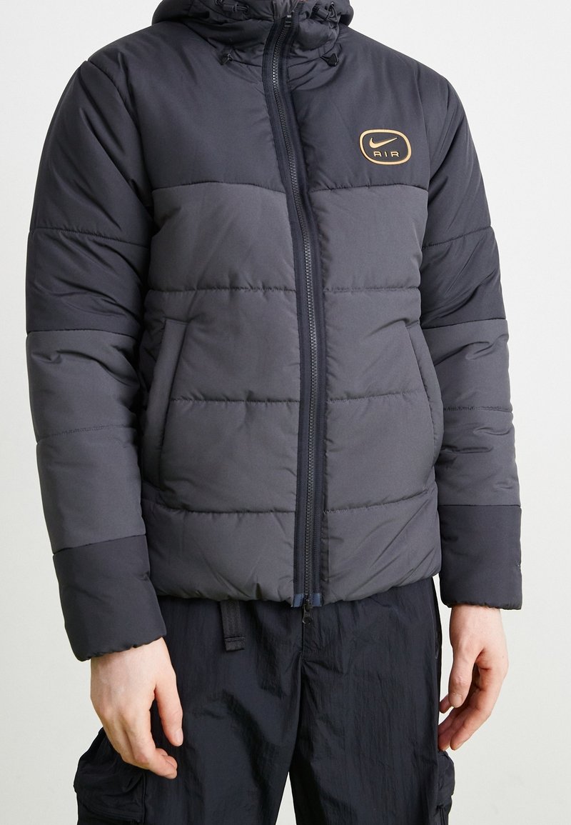 Svart och grå vadderad jacka med isolering och dragkedja, med en rund guldig Nike Air-logotyp på bröstet och huva. Texturerade paneler över hela plagget.