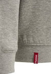 Grå sweatshirtmaterial med en melerad textur. Har en röd märkesetikett i nederkant, ribbade mudd och rundad halsringning.