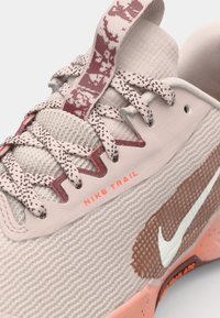 Nike Trail Sneaker mit einem strukturierten, hellrosa Mesh-Obermaterial, kontrastierendem weißen Logo, braunen Akzenten und gemusterten Schnürsenkeln. Mit einem gepolsterten Kragen.