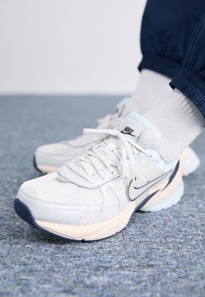 Białe sneakersy Nike noszone z białymi skarpetkami i granatowymi spodniami, stojące na niebieskim wykładanym dywanie podłożu.