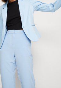 Jasnoniebieski żakiet wykonany z gładkiego materiału, z zapięciem na jeden guzik i dwiema bocznymi kieszeniami, w połączeniu z dopasowanymi jasnoniebieskimi spodniami.