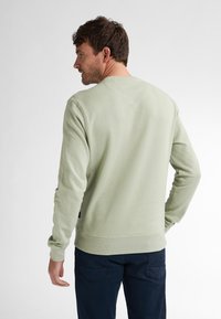 Sudadera verde claro con un acabado texturizado, cuello redondo y puños acanalados, llevada sobre un pantalón azul marino. Presenta sutiles motas y un diseño en la parte trasera.