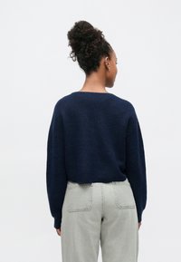 Pull bleu marine avec une finition texturée, manches larges et encolure arrondie, associé à un pantalon gris clair taille haute avec poches arrière.