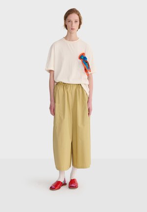 Junge Frau trägt lockere beige Hosen, ein übergroßes weißes T-Shirt mit blau-orangefarbenem Schal-Detail, weiße Socken und rote Sandalen mit offenen Zehen.