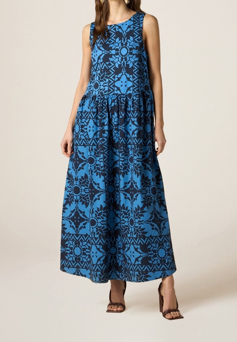 Robe maxi avec un motif floral bleu sur fond noir, sans manches, de forme fluide et taille froncée. Assortie à des sandales brunes à lanières.