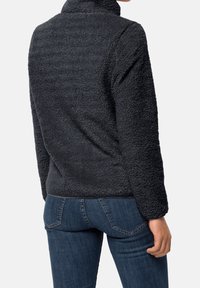 Osoba v černé fleecové bundě a modrých džínech, zobrazená zezadu, s jednou rukou uvolněnou podél těla.