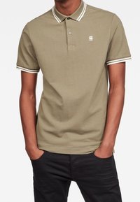 Polo beige avec col et poignets à rayures blanches, manches courtes, un petit logo sur la poitrine et une coupe classique. Texture en coton doux.