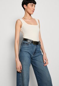 Débardeur ajusté beige clair avec un décolleté carré, associé à un jean bleu taille haute à jambes larges, featuring une ceinture noire avec une boucle dorée.