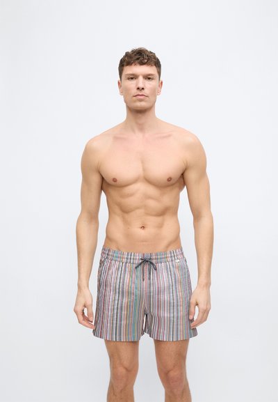 Νέος άνδρας με κοντά σγουρά μαλλιά στέκεται με πρόσωπο προς τα εμπρός, φορώντας πολύχρωμα μαγιό με κάθετες ρίγες και κορδονάκι στη μέση.