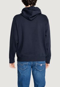 Felpa blu navy con cappuccio ampio, polsini e orlo a costine. Il tessuto è liscio e leggermente strutturato, abbinato a jeans in denim blu.