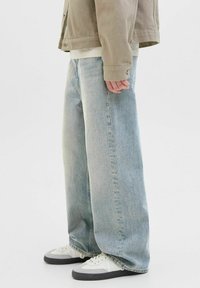 Jean large bleu clair en denim, avec taille haute et coutures minimales, associés à des baskets blanches et une veste beige.