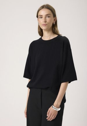 Black short-sleeve knit sweater with a round neckline and relaxed fit, paired with black trousers and a silver bangle on the wrist.
