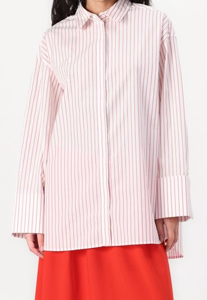 Frau trägt ein weißes, langärmeliges Hemd mit roten vertikalen Streifen und weiten Manschetten, kombiniert mit einem leuchtend roten Rock.