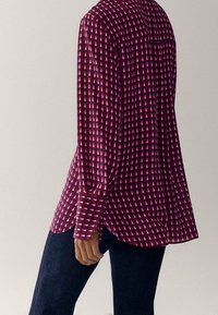 Femme portant une chemise à manches longues de couleur bordeaux avec des triangles géométriques blancs et un jean bleu foncé, se tenant devant un fond uni.