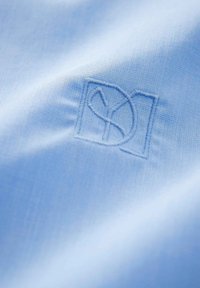 Un tessuto azzurro chiaro presenta una superficie leggermente texturizzata con un logo ricamato di blu scuro, posizionato all'interno di una cornice quadrata.