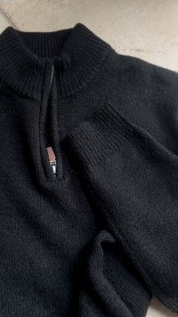 Black knitted sweater with a high collar and zip closure. Features ribbed cuffs and a soft, textured fabric. Distinctive leather zipper pull.