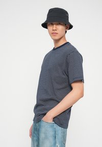 Νέος άνδρας με μαύρο καπέλο τύπου bucket, μπλούζα με ριγέ σκούρο μπλε και άσπρο, και ανοιχτό μπλε τζιν, με το ένα χέρι στην τσέπη, στέκεται σε λευκό φόντο.