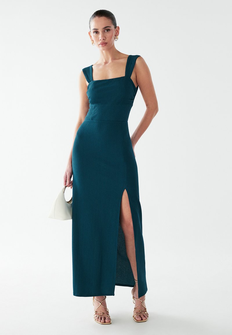 Teal golvlång klänning med fyrkantig urringning, strukturerat tyg, breda axelband och en slits i sidan, matchad med remsandaler och en vit handväska.