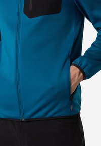 Giacca softshell blu con accentuazioni nere, frontale con zip e tasca, tessuto elasticizzato e polsini elastici. Texture liscia e design aderente.