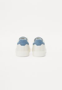 Baskets blanches avec un accent de talon en daim bleu, tige en cuir lisse, bout rond et semelle en caoutchouc texturé.