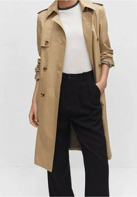 Beige trenchcoat met knoopaccenten, epauletten en een ceintuur in de taille, gecombineerd met een lichtgrijze blouse en zwarte high-waisted broek.