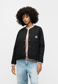 Giacca nera imbottita con bottoni a pressione, tasca sul petto e due tasche frontali. Indossata sopra un maglione rosa chiaro e abbinata a jeans azzurri.