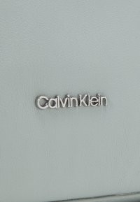 Svetlo siva usnjena površina s srebrnim logotipom "Calvin Klein". Tekstura je gladka s kamenčkasto površino.